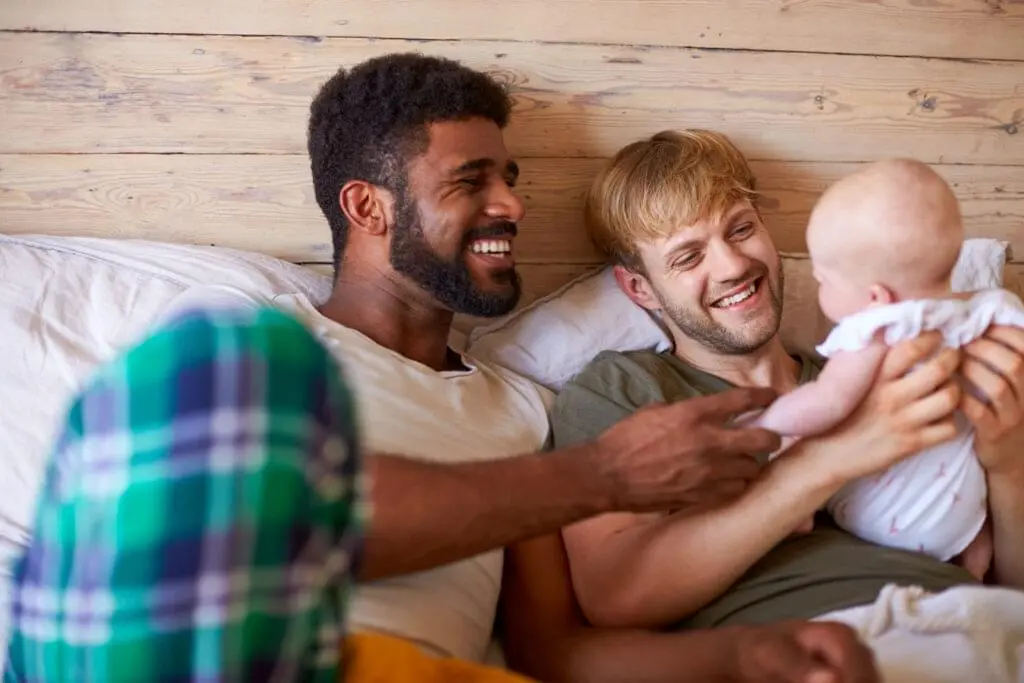 Loving Male Same Sex Couple Cuddling Baby Daughter In Bedroom At Home Together